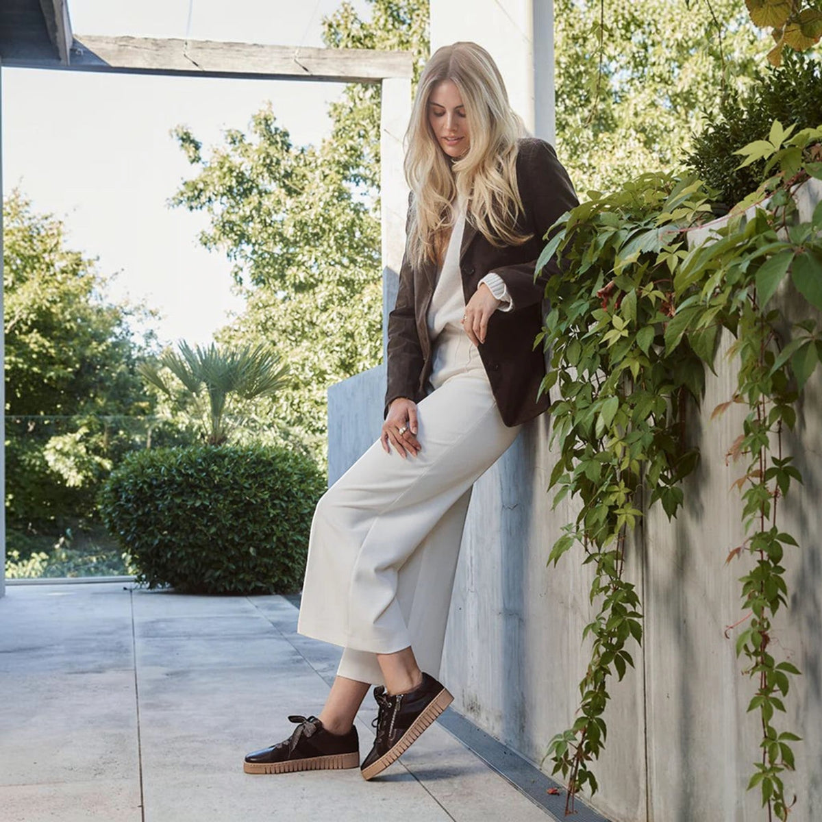 Woman wearing The Shoe Shop Tinah sneakers in dark brown leather with a platform sole in an outdoor garden setting.
