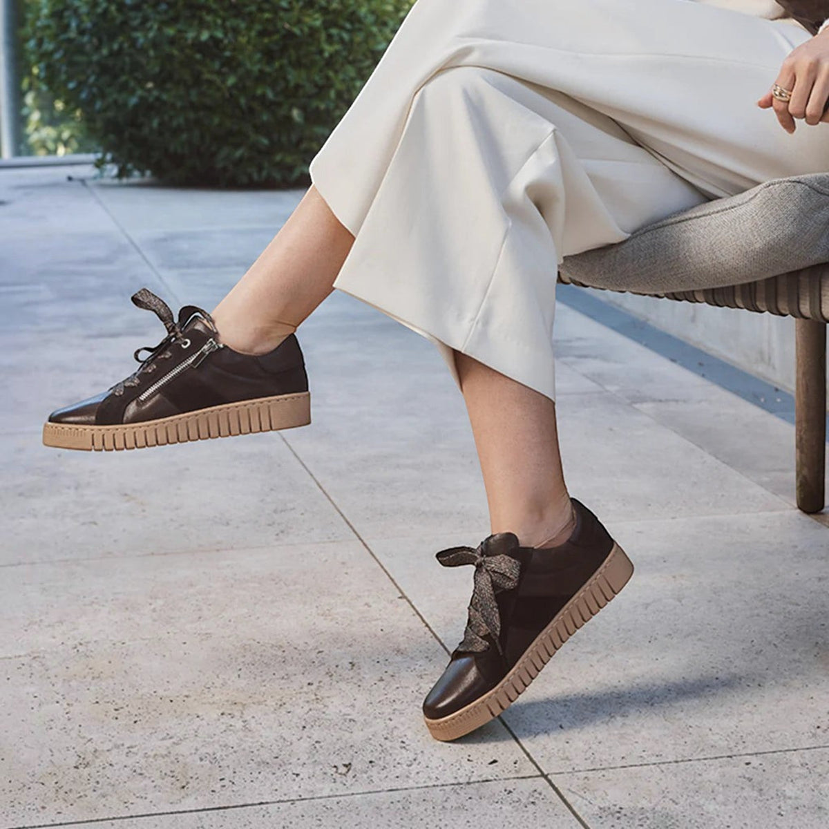 The Shoe Shop Tinah sneaker in dark brown leather with a side zipper and metallic ribbon laces on a seated model