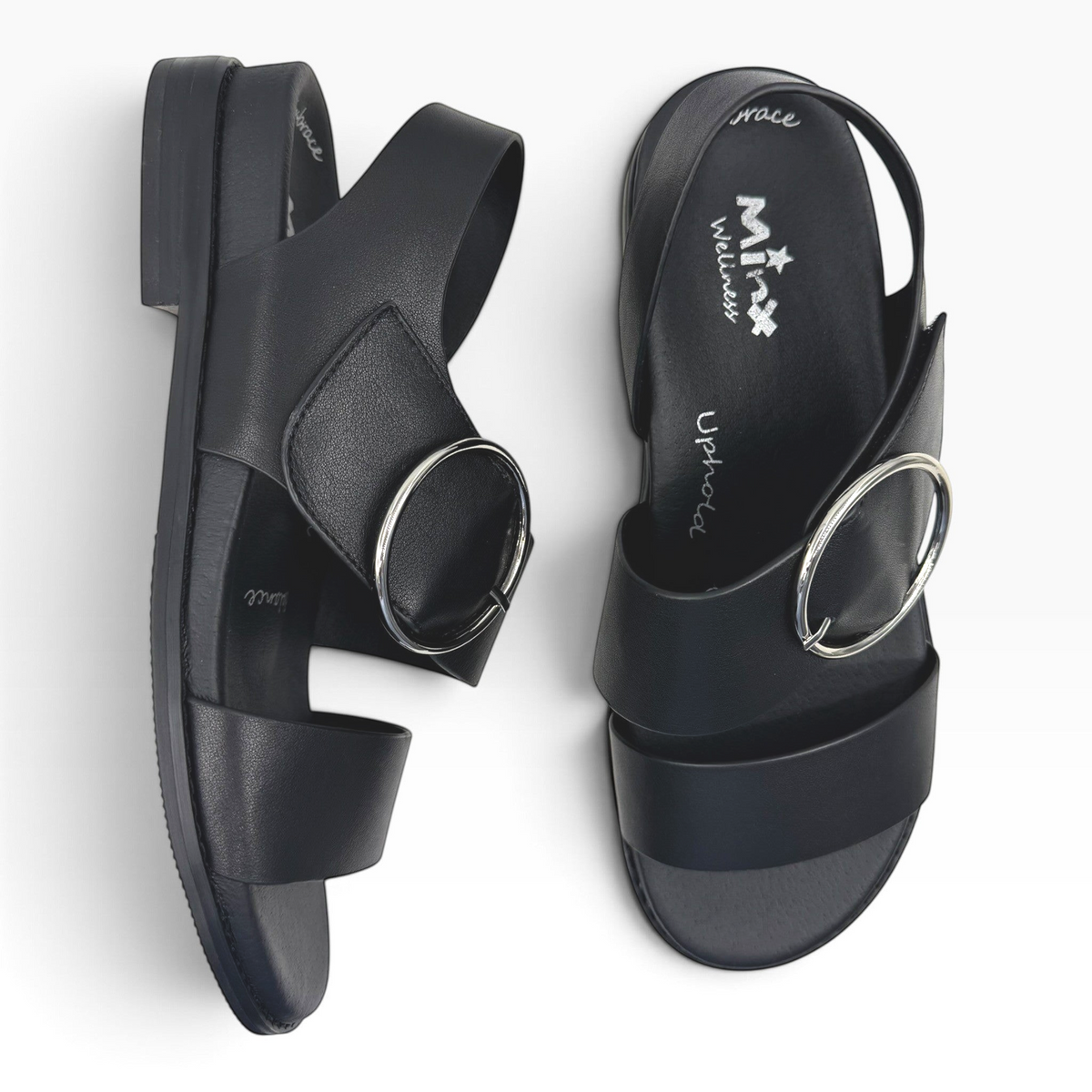The Shoe Shop Willa black leather flat sandals featuring a large silver circular buckle and adjustable ankle strap.